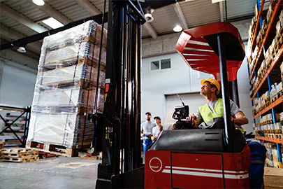 Ein Gabelstaplerfahrer mit gelbem Helm und zwei Lagerhelfer im Hintergrund arbeiten Hand in Hand in einem Logistikzentrum in Dresden, Sachsen.