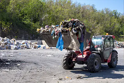 Ein roter Radlader mit einer riesigen, prall gefüllten Schaufel voll Mischmüll und Plastikabfällen fährt über das Außengelände eines Recyclinghofs in Dresden, Sachsen.