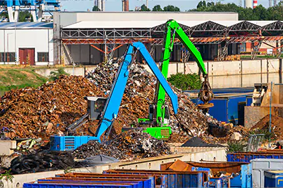 Ein großer Schrottplatz und Recyclinghof mit riesigen Bergen von Metallschrott, auf dem ein blauer und ein grüner Umschlagbagger mit Greifarm Altmetall sortieren und verladen.