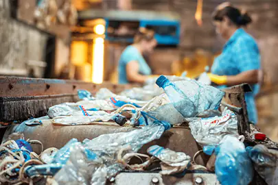 Zerdrückte Plastikflaschen und PET-Müll auf einem Förderband im Fokus, während im Hintergrund zwei Sortierkräfte in einer Recyclinganlage in Dresden, Sachsen, die manuelle Wertstofftrennung durchführen.